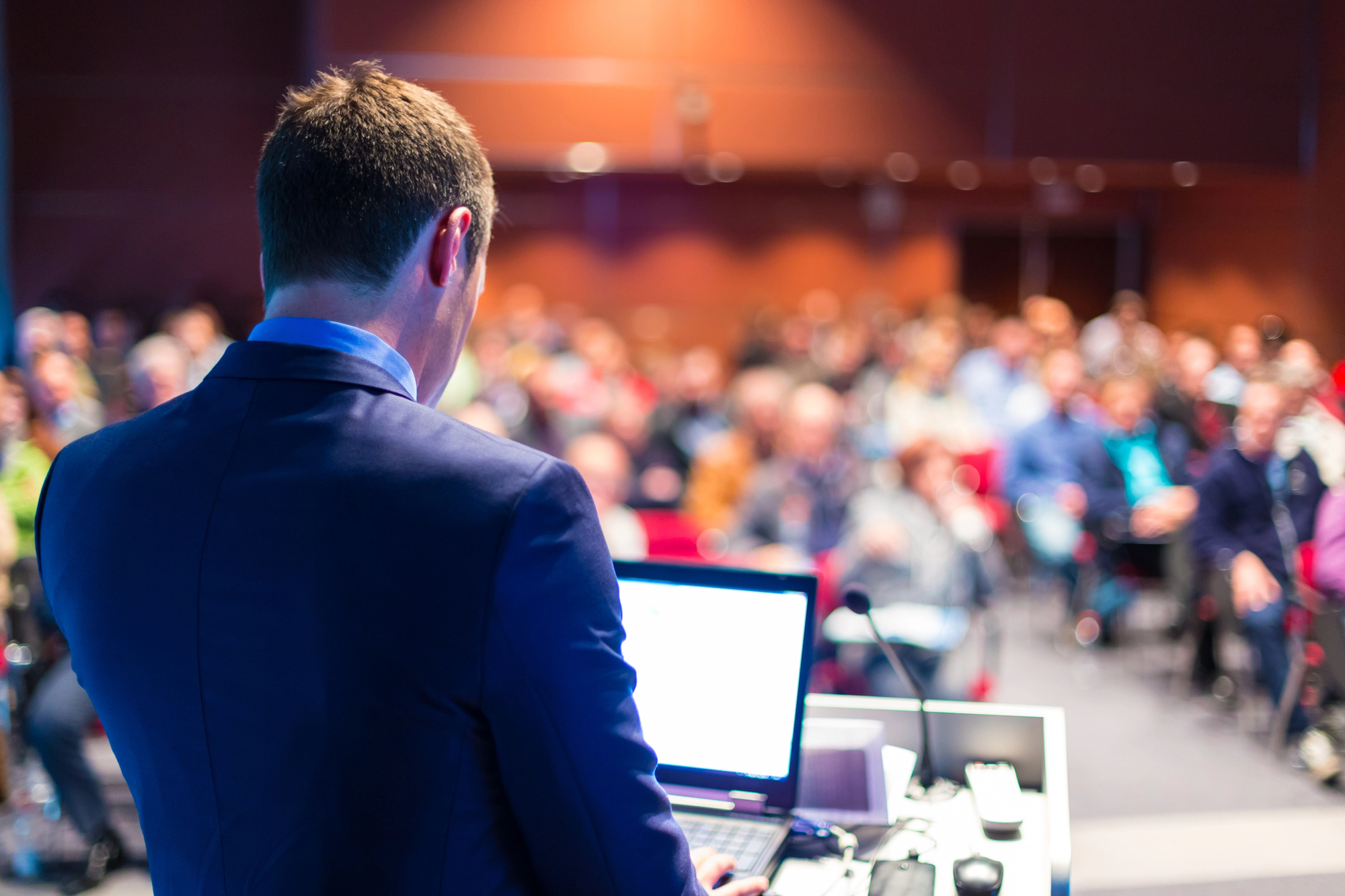 Man in a suit speaks at a podium with a laptop, addressing a large, blurred audience in a conference room—demonstrating how technology like Live Streaming for Wedding can connect distant guests to important events.