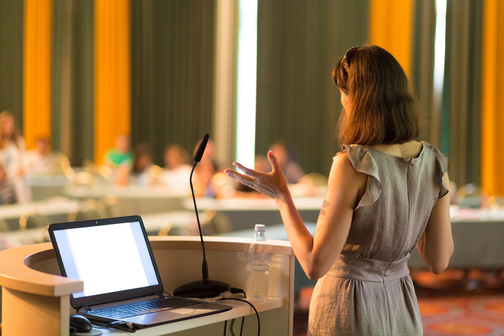A woman stands at a podium with a laptop and microphone, gesturing as she presents to a seated audience in a conference room, discussing innovative solutions such as Live Streaming for Wedding ceremonies.